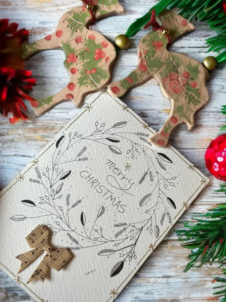 Festive gift bundle featuring a hand-illustrated Christmas card and two handmade pottery camel ornaments with holly berry designs, displayed among Christmas decorations