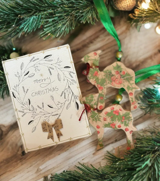 Flat lay of two handmade pottery camel ornaments with holly berry patterns beside a hand-illustrated ‘Merry Christmas’ card on textured paper, styled with festive greenery